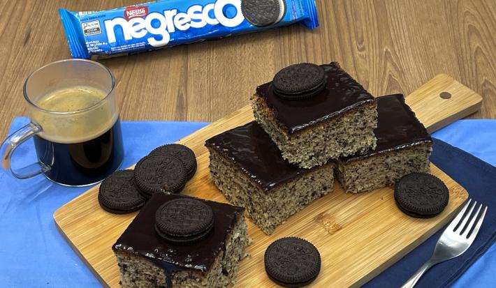 Fotografia em tons de azul com uma tábua de madeira ao centro. Em cima da tábua existe fatias de bolo formigueiro com cobertura de chocolate e biscoito negresco. Ao lado existe uma caneca de café.