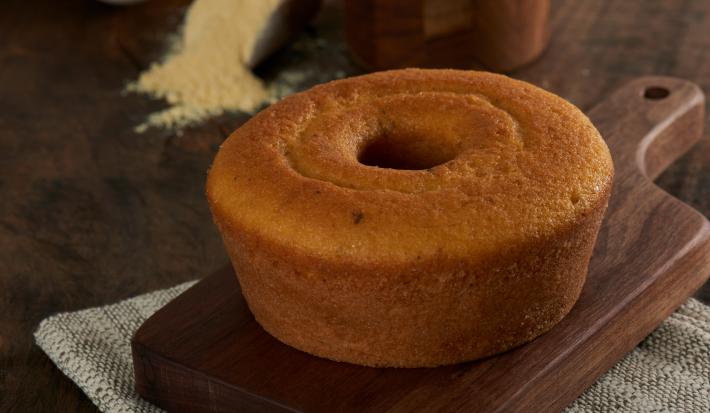 Foto aproximada do bolo de milho com erva-doce, redondo, com furo central, bem dourado, sobre uma tábua de madeira com um punhado de fubá aparecendo ao fundo
