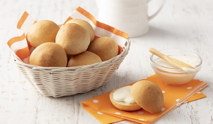 Fotografia em tons de laranja e branco de uma bancada branca com um paninho laranja, sobre ele um pãozinho aberto recheado e um recipiente de vidro com requeijão. Ao lado uma cesta com um paninho laranja e branco com os pães.