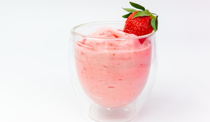 Fotografia de um creme de morango com erva cidreira em um recipiente de vidro de decoração sobre uma mesa branca. Em cima do creme, há um morango inteiro decorando.