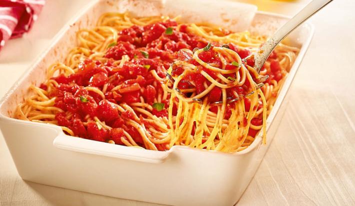 Fotografia em tons de marrom, amarelo e vermelho de uma bancada de madeira clara, sobre ela um recipiente quadrado branco com macarrão, molho sugo e uma colher de servir com uma porção do macarrão. Ao fundo um guardanapo vermelho com listras brancas.