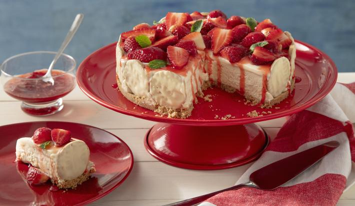 Fotografia em tons de vermelho em uma bancada de madeira de cor branca. Ao centro, uma boleira vermelha contendo a torta holandesa. Ao lado, há um pires com uma fatia da torta e um pano listrado vermelho.
