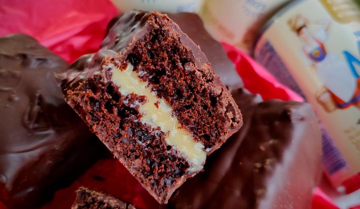 Imagem da receita de pão de mel com doce de leite moça, servido cortado ao meio mostrando o recheio, coberto com chocolate, sobre um tecido vermelho e com algumas latas de leite moça ao fundo