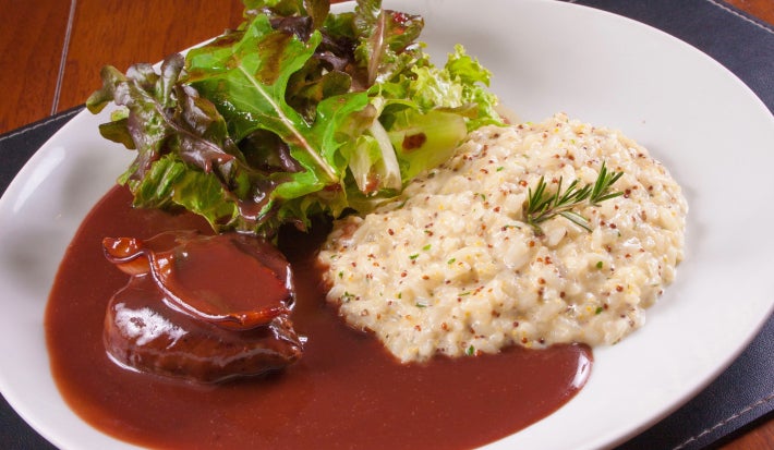 Fotografia em tons claros com uma receita de arroz a piamontese ao centro acompanhada de carne e folhas.