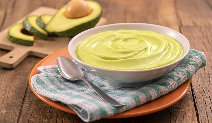 Foto em tons de verde da receita de creme rústico de abacate servida em um bowl de porcelana branco com uma colher prateada ao lado sobre um pano verde e branco quadriculado, tudo sobre um prato de porcelana laranja. Ao fundo, tábua com fatias de abacate