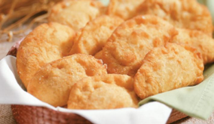 Fotografia em tons de verde em uma bancada de madeira com uma toalha de juta, uma cesta de vime marrom com guardanapo de pano verde e branco com pasteis de angu.
