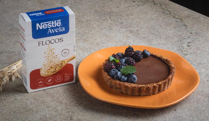 Foto da receita de Torta de Maçã com Chocolate e Nestlé Aveia Flocos, decorada com mirtilos, amoras e hortelã, servida em um prato laranja, sobre uma bancada de pedra onde está uma caixa de Nestlé Aveia Flocos e um ramo de trigo seco
