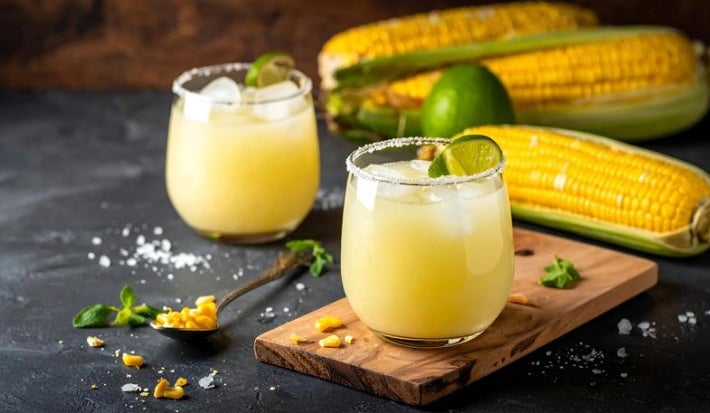 Fotografia de um copo de vidro sobre uma mini tábua de madeira retangular, sobre uma mesa preta. Ao fundo tem outro copo e três espigas de milho. Dentro dos copos tem o suco de milho, menos cremoso.