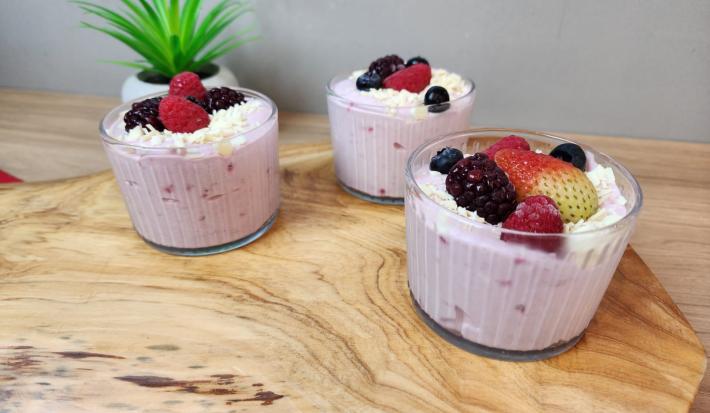 Fotografia em tons de rosa em uma bancada de madeira clara com três potes de vidro e mousse dentro de cada um deles. Frutas vermelhas decorando.