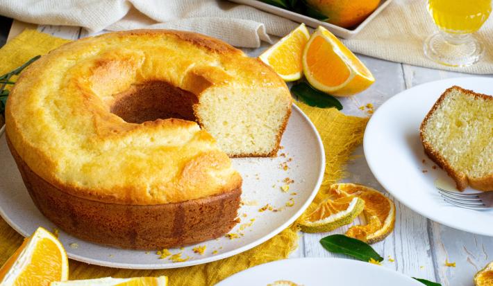 Fotografia de um bolo de laranja feito em forma de furo central sobre um prato raso de cerâmica branco. Ao lado tem um prato de sobremesa com uma fatia do bolo e um garfo, e ao redor tem pedaços e fatias de laranja, sobre uma mesa de madeira branca.