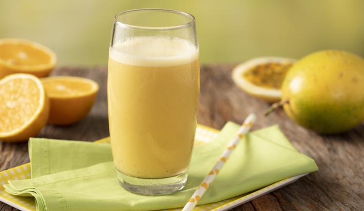 Fotografia em tons de amarelo, verde e marrom de uma bancada de madeira com uma bandeja amarela e um paninho verde com um canudo sobre ele um copo de vidro com a vitamina de laranja. Ao fundo três metades de laranjas, um maracujá inteiro e uma metade.