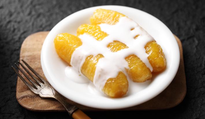 Fotografia de três bananas carameladas com creme de leite por cima.  As bananas estão em um prato pequeno branco raso, ao lado de um garfo que está apoiado em uma tábua de madeira pequena.