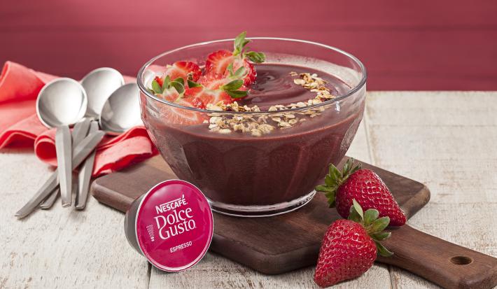 fotografia em tons de marrom e vermelho vista de uma bancada cinza, contém uma tábua de madeira com um recipiente transparente com o açaí e morangos por cima para decorar. Uma capsula de Dolce Gusto e morangos na frente e ao fundo colheres.