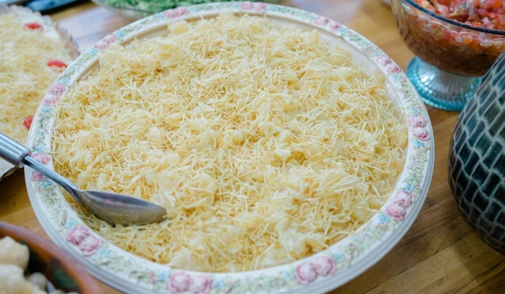 Fotografia de um prato grande redondo com salada de batata coberta com batata-palha crocantes, decorado com flores. Ao fundo, uma salada verde com pedaços de fruta amarela.
