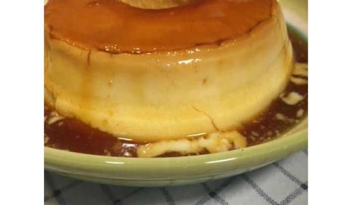 Fotografia em tons de bege em uma bancada com um prato redondo bege com detalhes verde e o pudim de leite inteiro em cima dele
