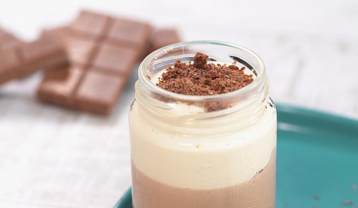 Fotografia em tons de branco e azul de uma bancada branca com um prato retangular azul, sobre ele um copo de vidro com o cheesecake. Ao fundo tabletes de chocolate.