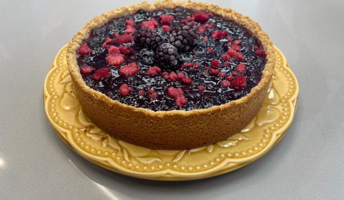 Foto da receita de torta ridícula servida em uma porção grande decorada com frutas vermelhas em cima. Torta está sobre um prato amarelo em cima de uma bancada cinza