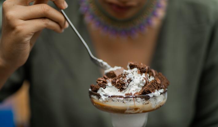 Fotografia de uma pessoa segurando uma colher de sopa e comendo uma sobremesa de camadas de massa e MOÇA Leite Condensado Cremoso. O doce está em uma tacinha de vidro.