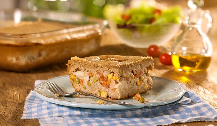 Fotografia em tons de marrom em uma bancada de madeira com um pano xadrez azul e um prato branco com um pedaço de torta integral de legumes. Ao fundo, uma travessa com a torta inteira, um potinho com salada e um vidro de azeite.