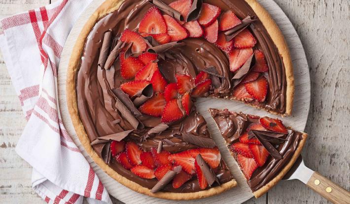 Fotografia em tons de branco e vermelho de uma bancada de madeira branca com uma tabua redonda com a pizza de chocolate com morangos. Ao lado um paninho branco com listras vermelhas e uma espátula retirando uma fatia da pizza.