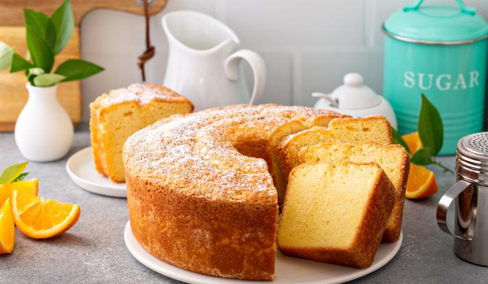 Fotografia de um bolo de laranja feito em forma redonda com furo no meio, o bolo está sobre um apoio, e tem três fatias cortadas, uma apoiada na outra. Ao fundo tem uma fatia do bolo em um prato de vidro pequeno, ao lado de pedaços de laranja.