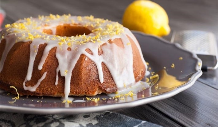 Fotografia de um bolo de limão em uma elegante travessa, coberto com glacê branco que escorre pelas laterais. Raspas de limão decoram o topo, enquanto um limão inteiro e um ralador estão ao fundo.