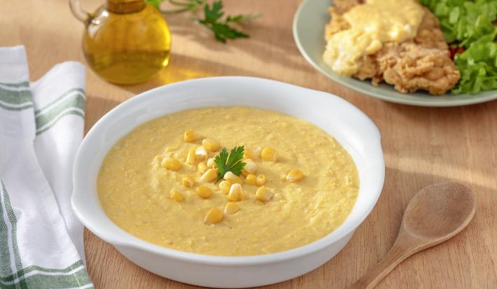 Foto da Receita de Creme de Milho Sem Lactose. Observa-se um recipiente branco com o creme de milho dentro e, atrás, um prato de filé à milanesa servido com o creme