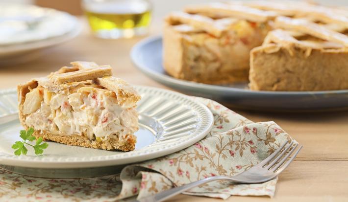 Fotografia em tons de bege em uma mesa de madeira com um pano florido em bege e vermelho e um prato branco apoiado nele com um pedaço de torta de palmito e alho-poró. Ao fundo, um prato grande com a torta cortada ao meio.