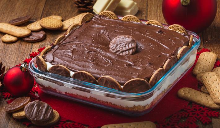 Foto da receita de Torta Holandesa na Travessa, em uma bancada de madeira decorada com temas de natal, alguns biscoitos espalhados, pedaços de chocolate branco. A receita tem um creme marrom sobre um creme claro e está em um refratário de vidro Foto da receita de Torta Holandesa na Travessa, em uma bancada de madeira decorada com temas de natal, alguns biscoitos espalhados, pedaços de chocolate branco. A receita tem um creme marrom sobre um creme claro e está em um refratário de vidro