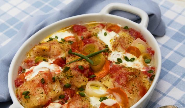 Um pano xadrez nas cores azul e branco, acima um recipiente contém o peixe com legumes e ao lado uma colher.