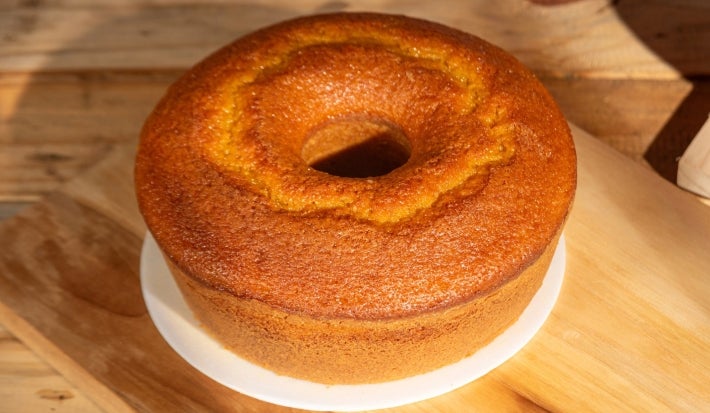 Um bolo de cenoura simples, com furo no meio, assado e com a superfície dourada e brilhante. Ele está sobre uma tábua de madeira clara, com um fundo de madeira natural, iluminado pela luz do sol.