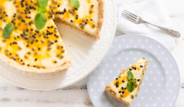 fotografia em tons de branco e amarelo de uma bancada branca vista de cima. Contém um prato branco redondo com uma torta com mousse de maracujá e sementes para decorar, e ao lado um prato redondo azul com a fatia da torta.