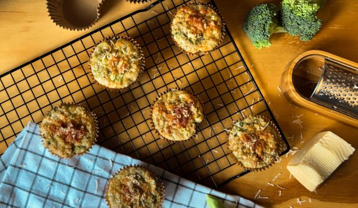 Fotografia vista de cima de uma mesa amarela com pedaços de brócolis cru, ralador, um pedaço de queijo e algumas forminhas de tom marrom. Sobre a mesa tem também uma grade preta e um pano azul, apoiado na grande tem seis muffins integrais de brócolis