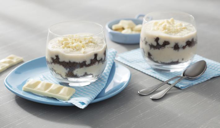 Fotografia em tons de azul em uma bancada de madeira cinza, um pano azul, um prato redondo azul claro e duas taças de vidro com o pavê de chocolate ao leite com chocolate branco, Galak. Ao fundo, um potinho com quadradinhos de chocolate Galak.