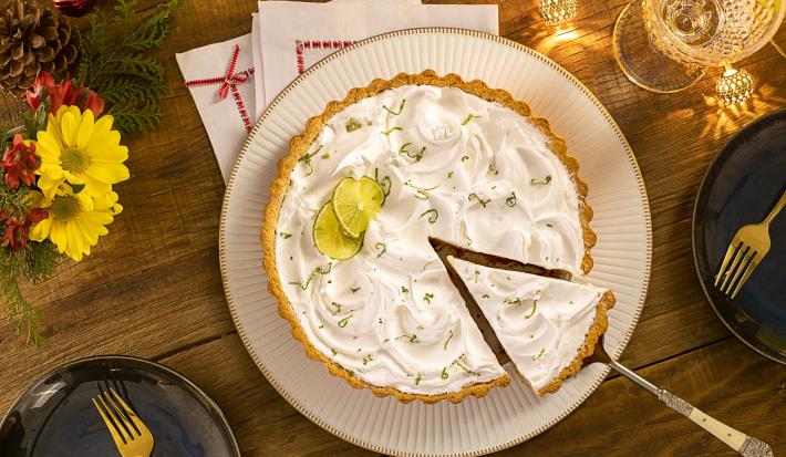 Imagem vista de cima de uma torta de limão com merengue e uma fatia sendo retirada. Ao redor, decoração de Natal.