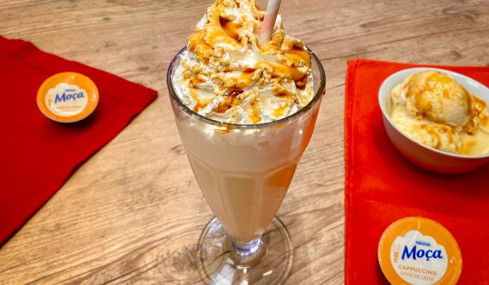 Fotografia em tons de amarelo em uma bancada de madeira de cor marrom. Ao centro, um copo contendo o milkshake. Ao lado, há dois panos vermelhos estendidos- em um lado está a cápsula de dolce gusto e do outro lado também há uma cápsula e um sorvete.