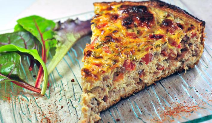 Fotografia mostra uma fatia de torta com camadas bem definidas de massa leve e recheio colorido de sardinha, tomate e azeitonas, polvilhada com orégano.