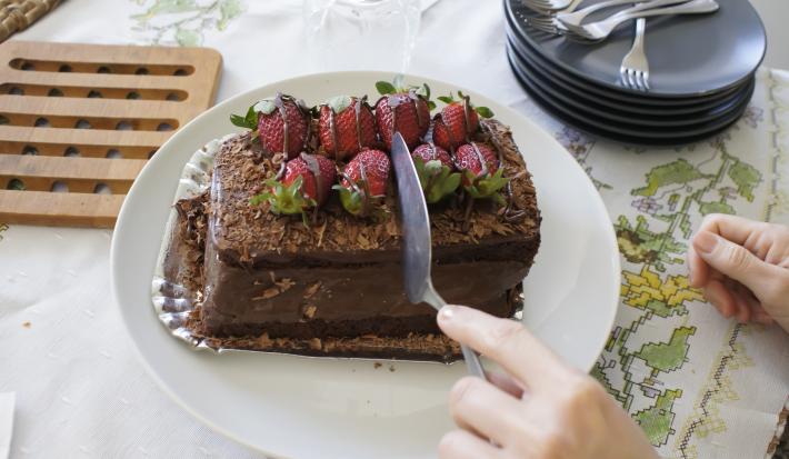 Fotografia de uma mão cortando um bolo de chocolate recheado com morango que está em um prato branco. Ao fundo, alguns pratos pretos empilhados, e ao lado, uma decoração em madeira para apoiar bolo ou pães. Tudo está sobre uma toalha de mesa branca.