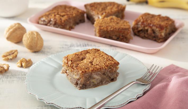 Foto da Receita de Bolo de Banana, Leite Moça e Nozes Caramelizadas. Observa-se uma fatia de foto no plano principal com um prato de sobremesa azul e decorados com banana e nozes.