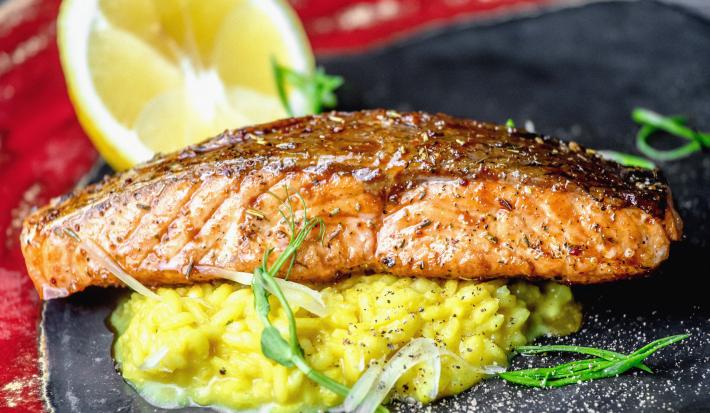 Fotografia de um filé de salmão grande sobre um risoto de limão sicilinao em um prato preto raso, salpicado com cebolinha. No prato tem metade de um limão siciliano.