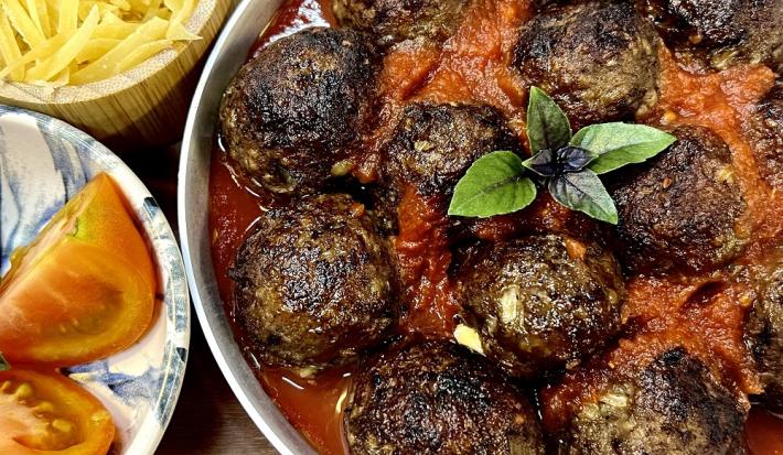 Foto em tons de vermelho escuro da receita de almôndegas recheadas servida em um prato grande e fundo de porcelana branco. Ao lado dois pratos menores, um com tomates e outro com queijo ralado.