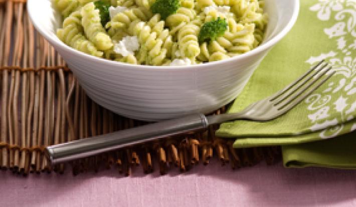 Fotografia em tons de verde em uma mesa de madeira com uma toalha rosa e um jogo americano de palha marrom.  Ao centro, um prato branco fundo redondo com o macarrão tipo parafuso com o molho pesto de brócolis e ao lado um guardanapo de pano verde.