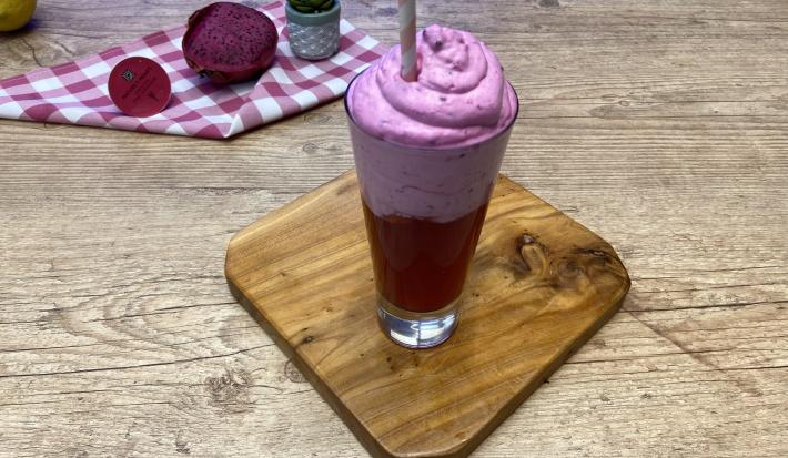 Fotografia em tons de rosa em uma bancada de madeira clara com uma tábua de madeira ao centro e um copo de vidro alto com uma bebida rosa e uma espuma rosa. Ao fundo, um pano xadrez rosa com uma pitaya aberta na metade e um limão-siciliano.