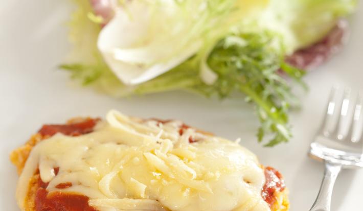 Fotografia em tons de marrom em uma bancada de madeira com uma toalha cinza, um sousplat marrom, um prato branco redondo com o filé à parmegiana acompanhado de salada. Ao fundo, um potinho de azeite.
