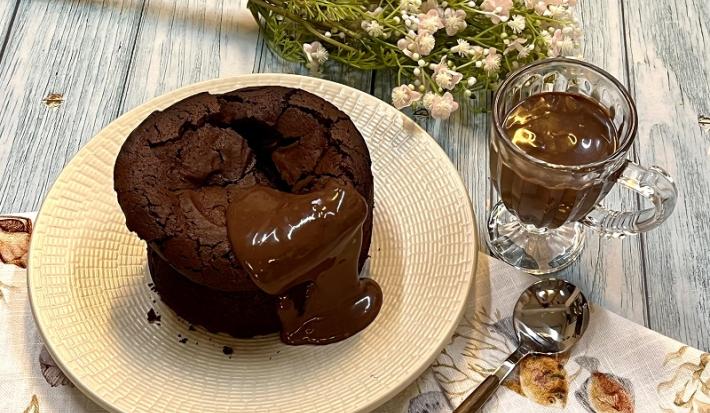 Foto em tons de bege e marrom da receita de bolo fudge servida sobre um prato bege e uma tolha de mesa decorada. Ao lado, há uma colher prateada e uma caneca de vidro com chocolate derretido. Ao fundo, um buqué de flores cor de rosa
