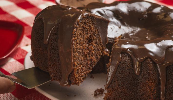 Foto bem aproximada de um bolo de chocolate com uma fatia sendo retirada. O bolo está num prato branco e coberto com Brigadeiro e o prato está sobre uma toalha xadrez vermelho e branco.