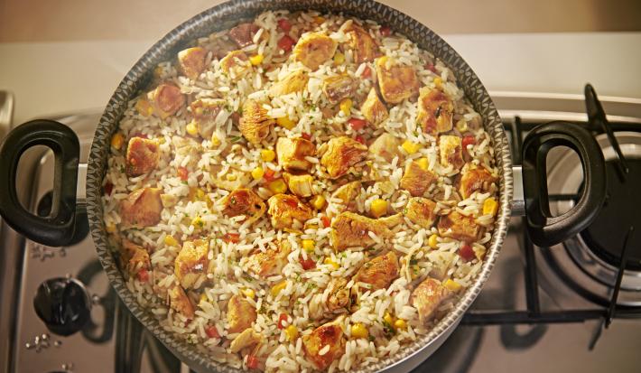 Foto de cima de uma panela com a receita de arroz caipira, em tons branco e amarelo, com pedaços de frango e milho. A panela está sobre um fogão e dela sai uma fumaça.