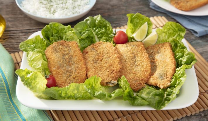 Fotografia em tons de verde em uma bancada de madeira, um pano verde com azul, um jogo americano de madeira clara, um prato branco redondo com a sardinha frita acompanhada de salada de alface e tomate cereja. Ao fundo, um potinho com azeite.