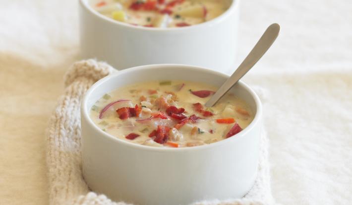Fotografia de dois recipientes de sopa fundos e na cor branca, com um caldo de batata e salsão com cenoura, bacon, cebola roxa e massa. O recipiente da frente está apoiado em uma toalha de lá na cor bege, que está sobre uma mesa na cor bege claro.
