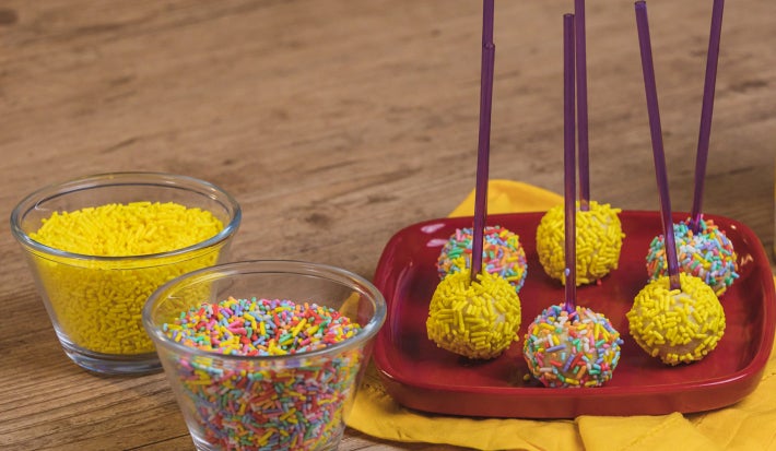 Fotografia em tons de amarelo e vermelho em uma bancada de madeira clara, um pano amarelo e um prato vermelho com alguns pirulitos de ninho com granulado colorido em cima dele. Ao lado, potinhos com os granulados coloridos e a lata de Ninho em Pó.